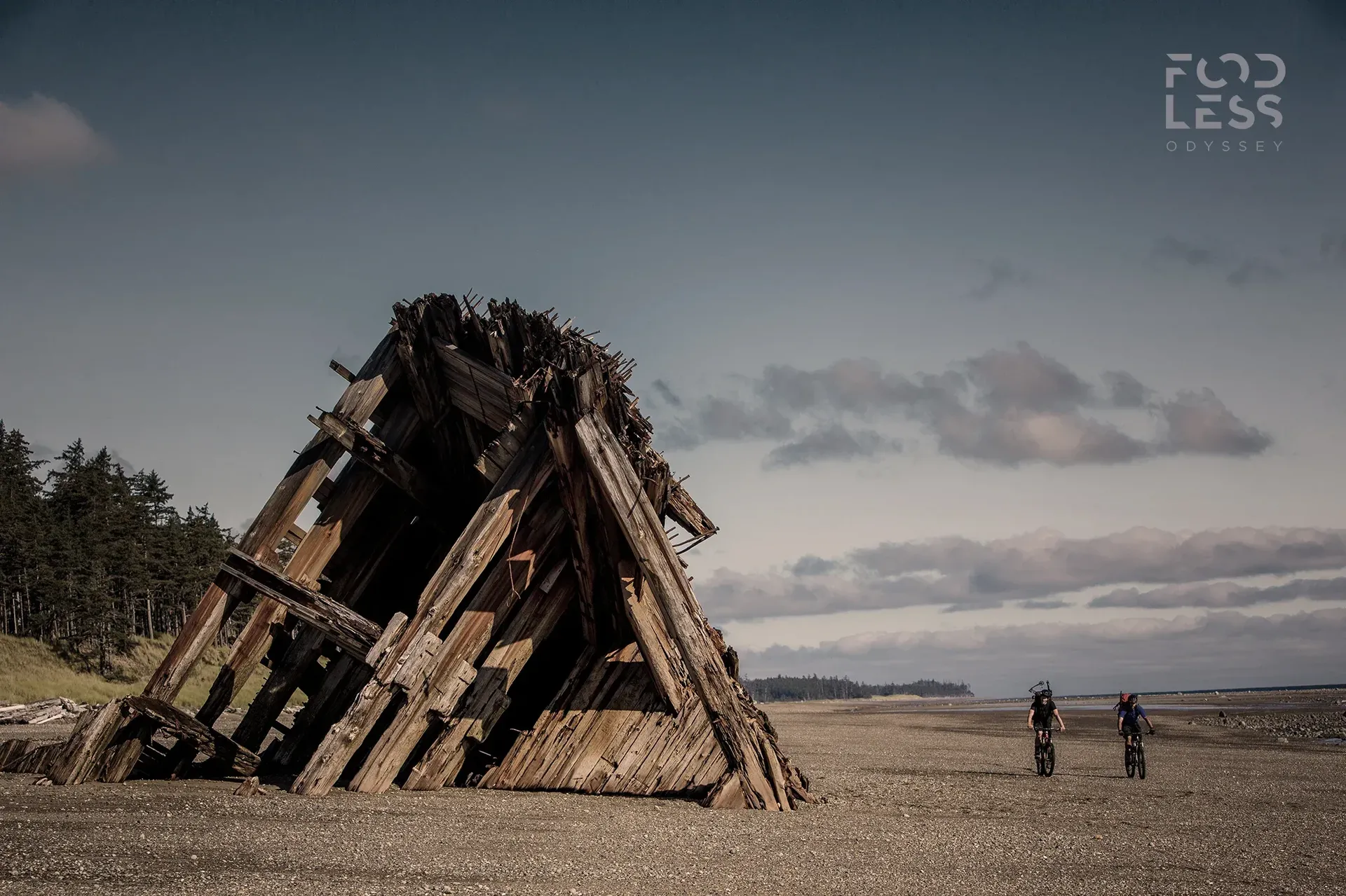 2 cyclists ride past a wooden beach shelter
